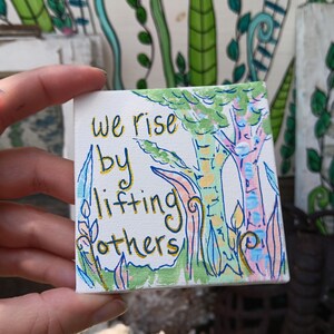 May include: A small square canvas painting featuring the hand-lettered text "we rise by lifting others" in gold ink. The artwork depicts a stylized forest scene with green, pink, and blue trees and foliage painted in a light, colorful palette.