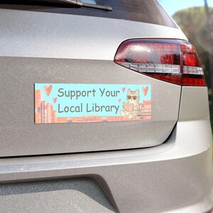 Könnte beinhalten: Ein rechteckiger Autoaufkleber mit hellblauem Hintergrund und dem Text "Unterstützen Sie Ihre örtliche Bibliothek". Der Aufkleber zeigt eine Cartoon-Katze mit Sonnenbrille, die ein Buch liest, mit Herzen und Bücherstapeln.