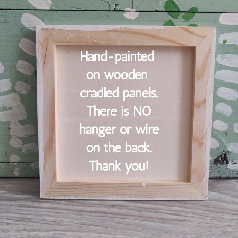 May include: A square, wooden-framed canvas with the text "Hand-painted on wooden cradled panels. There is NO hanger or wire on the back. Thank you!" The frame is made of light-colored wood.