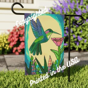 以下が含まれることがあります： ピンクの花の近くにホバリングしている、長い嘴を持つ緑、青、紫のハチドリが描かれたカラフルなガーデンフラッグ。フラッグは黄色い背景に緑の葉と青と緑の渦巻き模様。フラッグの上部には白い文字で「Copyrighted」、下部には白い文字で「Printed in the USA」と印刷されています。
