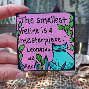 May include: A small square canvas painting with a pink background featuring a teal cat illustration and green leaf accents. The text reads: The smallest feline is a masterpiece. Leonardo da Vinci. The painting has a black border and is held by fingers.