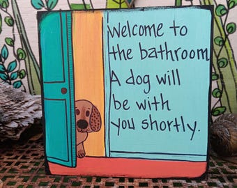May include: A square wooden sign painted in light blue, orange, and yellow, featuring a cartoon dog peeking from behind a door. The text reads: Welcome to the bathroom A dog will be with you shortly. The sign has a black border and hand-painted details.