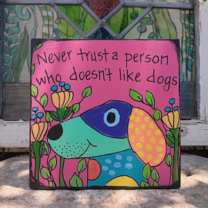 May include: A square, hand-painted wooden sign with a pink background and the text "Never trust a person who doesn't like dogs." The sign features a colorful cartoon dog with a blue, green, and yellow design, surrounded by flowers.
