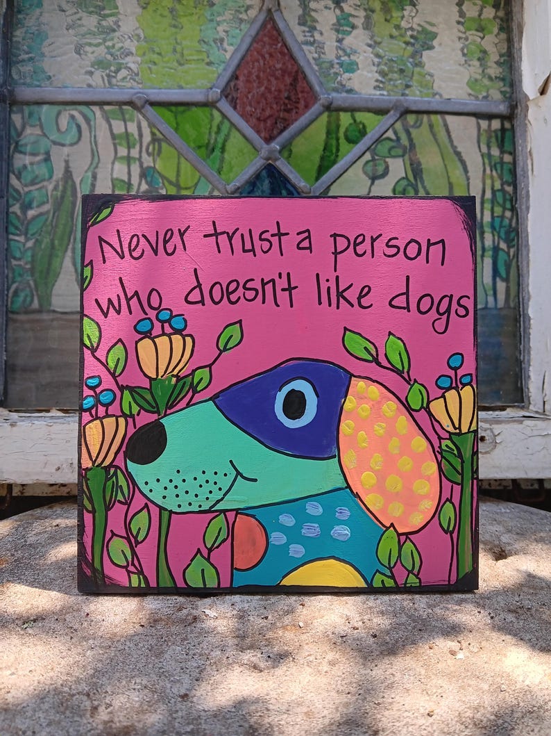 May include: A square, hand-painted wooden sign with a pink background and the text "Never trust a person who doesn't like dogs." The sign features a colorful cartoon dog with a blue, green, and yellow design, surrounded by flowers.