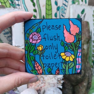 May include: A square sign with a bright blue background featuring hand-drawn black text that reads please flush only toilet paper. The sign is decorated with colorful illustrations of pink, yellow, and orange flowers with green leaves and stems.