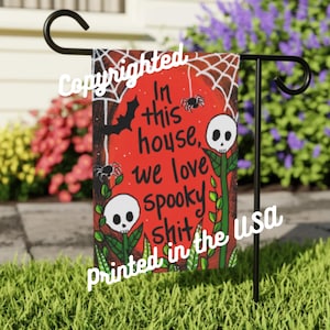 Puede incluir: Una bandera de jardín de Halloween roja y negra con una telaraña, murciélagos y calaveras. El texto en la bandera dice "In this house, we love spooky shit. Printed in the USA."