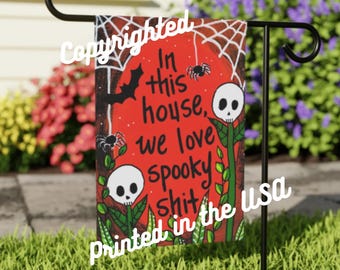 May include: A red and black Halloween garden flag with a spiderweb, bats, and skulls. The text on the flag reads "In this house, we love spooky shit. Printed in the USA."