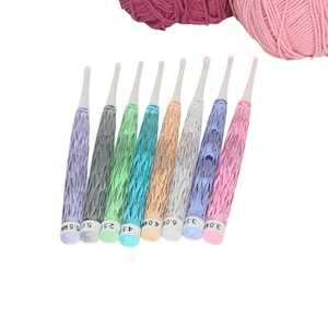 May include: A set of eight crochet hooks with textured, translucent handles in various colors including purple, gray, green, blue, orange, silver, and pink, positioned below two balls of yarn in dark pink and light pink shades.