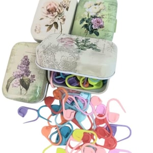 May include: Three rectangular metal tins with floral designs in cream, green, and light green, featuring vintage-style botanical illustrations. One tin is open, revealing a collection of assorted plastic stitch markers in pastel colors.