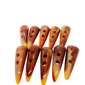 May include: A collection of ten elongated toggle buttons arranged in two rows. Each button features a smooth, polished surface with a marbled pattern in shades of amber, brown, and cream, and includes two circular holes for attachment.