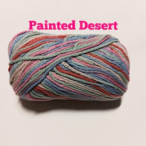May include: A skein of variegated yarn featuring a multi-colored palette of soft pink, sage green, light blue, and tan tones. The text "Painted Desert" is printed in bright pink letters above the yarn, which is wound into a compact oval shape.