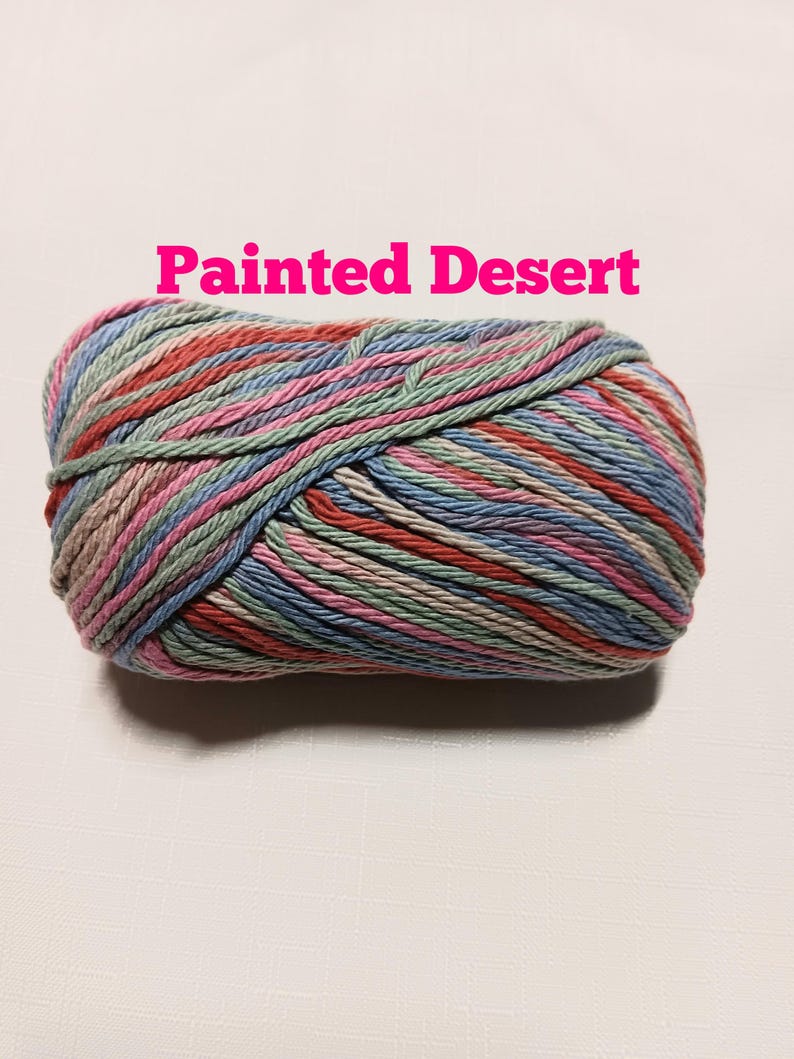 May include: A skein of variegated yarn featuring a multi-colored palette of soft pink, sage green, light blue, and tan tones. The text "Painted Desert" is printed in bright pink letters above the yarn, which is wound into a compact oval shape.
