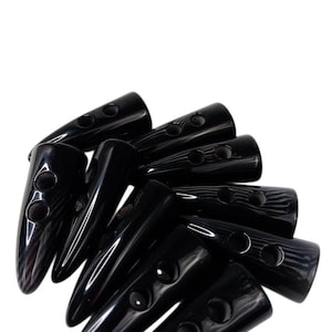 May include: A collection of black toggle buttons, each featuring a smooth, glossy finish and a tapered, elongated shape. Every button includes two circular holes positioned along the center for attachment, arranged in a scattered, overlapping pile.