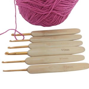 May include: A set of six crochet hooks with gold-colored metal tips and light-colored wooden handles, each marked with sizes 2.5mm, 3.0mm, 4.0mm, 4.5mm, 5.0mm, and 5.5mm, positioned below a large ball of vibrant pink yarn with a loose strand.