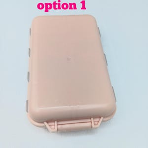 May include: A rectangular, light pink plastic storage case with rounded corners and a hinged lid. The text "option 1" is printed in pink above the case. The container features a secure latch mechanism on the front edge and visible side hinges.