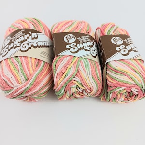May include: Three skeins of Lily Sugar'n Cream yarn in a pastel color palette of pink, green, yellow, and white. Each skein is wrapped with a brown label. The yarn is ideal for crochet and knitting projects.