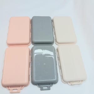 May include: Six rectangular plastic storage cases arranged in two rows of three. The top row features one light pink, one gray, and one beige case. The bottom row contains one light pink, one gray, and one beige case, each with a small latch.