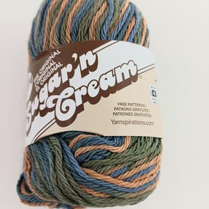May include: A skein of Sugar'n Cream yarn featuring a variegated color palette of sage green, dusty blue, and tan. The yarn is wrapped in a paper label with the text Sugar'n Cream, The Original, and Yarnspirations.com, indicating a medium 4 weight.