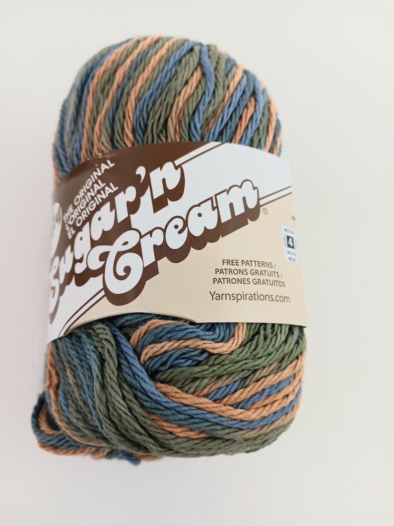 May include: A skein of Sugar'n Cream yarn featuring a variegated color palette of sage green, dusty blue, and tan. The yarn is wrapped in a paper label with the text Sugar'n Cream, The Original, and Yarnspirations.com, indicating a medium 4 weight.