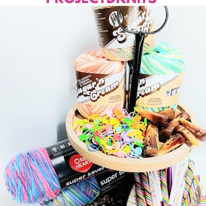 May include: A tiered wooden tray holding Sugar'n Cream yarn skeins, colorful plastic stitch markers, and brown resin toggles. Below, Red Heart Super Saver yarn in pink and blue, a collection of bee-shaped buttons, and rainbow-striped crochet hooks.