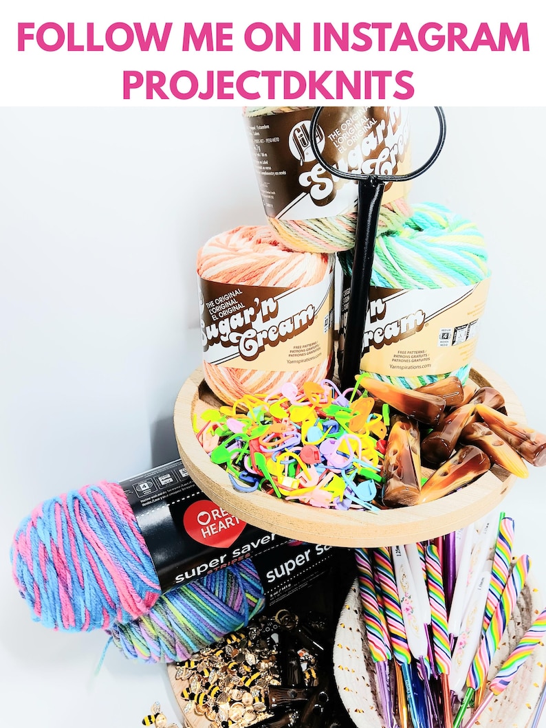 May include: A tiered wooden tray holding Sugar'n Cream yarn skeins, colorful plastic stitch markers, and brown resin toggles. Below, Red Heart Super Saver yarn in pink and blue, a collection of bee-shaped buttons, and rainbow-striped crochet hooks.