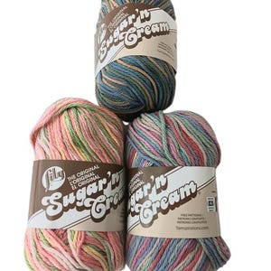 May include: Three skeins of Lily Sugar'n Cream yarn arranged in a triangle. The top skein features blue, green, and tan variegated tones. The bottom left skein is pink, green, and yellow, while the bottom right is blue, pink, and green.