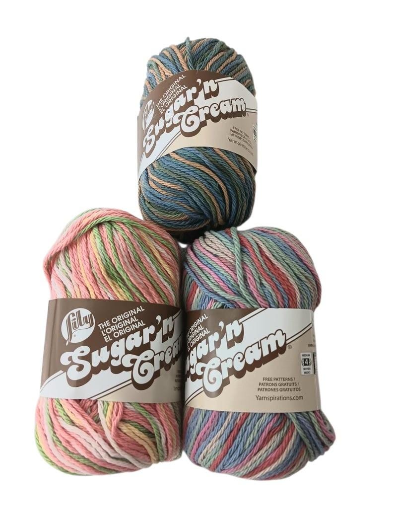 May include: Three skeins of Lily Sugar'n Cream yarn arranged in a triangle. The top skein features blue, green, and tan variegated tones. The bottom left skein is pink, green, and yellow, while the bottom right is blue, pink, and green.