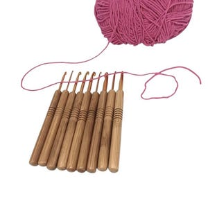 May include: A set of nine crochet hooks with light brown bamboo handles featuring horizontal grooved detailing and metallic tips, arranged in a row alongside a single ball of vibrant pink yarn with a strand extending across the hook tips.
