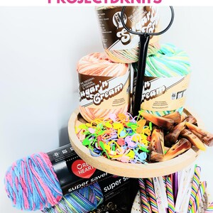 May include: A tiered wooden tray holding Sugar'n Cream yarn skeins, colorful plastic stitch markers, and brown resin toggles. Below, Red Heart Super Saver yarn in pink and blue, a collection of bee-shaped buttons, and rainbow-striped crochet hooks.