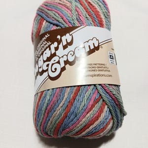 May include: A skein of Sugar'n Cream yarn featuring a variegated color palette of muted pink, sage green, and dusty blue. The paper label displays the brand name, the text "FREE PATTERNS," and a medium weight 4 symbol on a tan background.