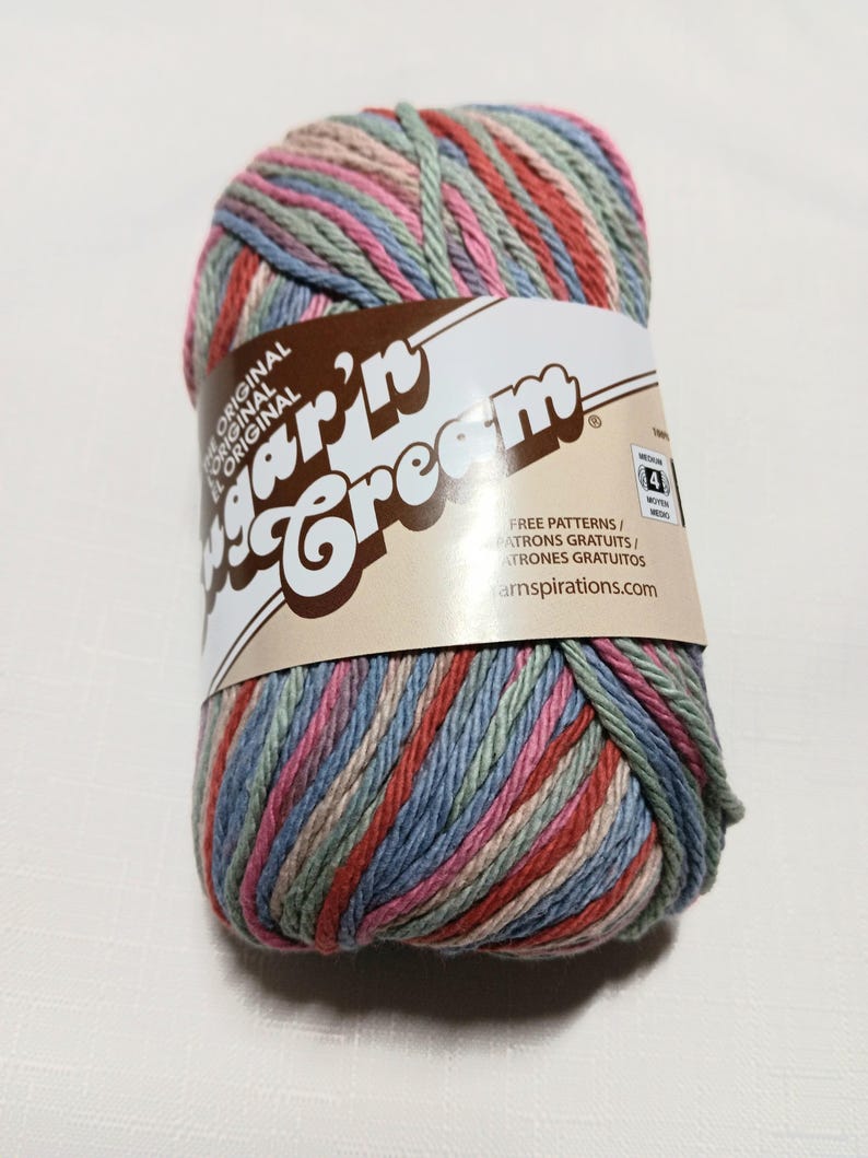 May include: A skein of Sugar'n Cream yarn featuring a variegated color palette of muted pink, sage green, and dusty blue. The paper label displays the brand name, the text "FREE PATTERNS," and a medium weight 4 symbol on a tan background.