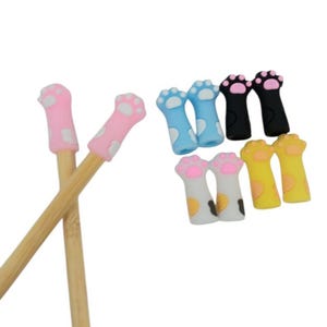 May include: Two bamboo knitting needles topped with pink silicone cat paw caps, accompanied by eight additional silicone cat paw caps in blue, black, white with brown spots, and yellow, all featuring raised paw pads and toe details.
