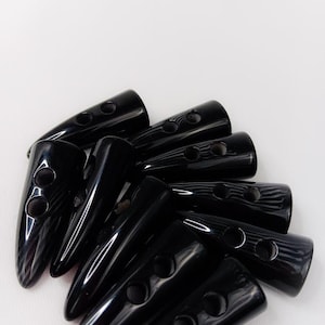 May include: A collection of glossy black toggle buttons. Each button is elongated with a rounded end and two holes for sewing. The buttons are arranged against a white background, showcasing their smooth, reflective surface.