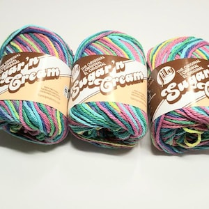 May include: Three skeins of Lily Sugar'n Cream yarn in a vibrant mix of colors including pink, blue, green, and yellow. Each skein is wrapped with a brown label featuring the brand name and logo.