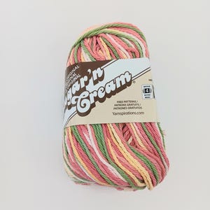 May include: A skein of Sugar'n Cream yarn featuring a variegated color palette of pink, green, yellow, and white strands. The paper label displays the brand name, a medium weight 4 symbol, and the website Yarnspirations.com for free patterns.