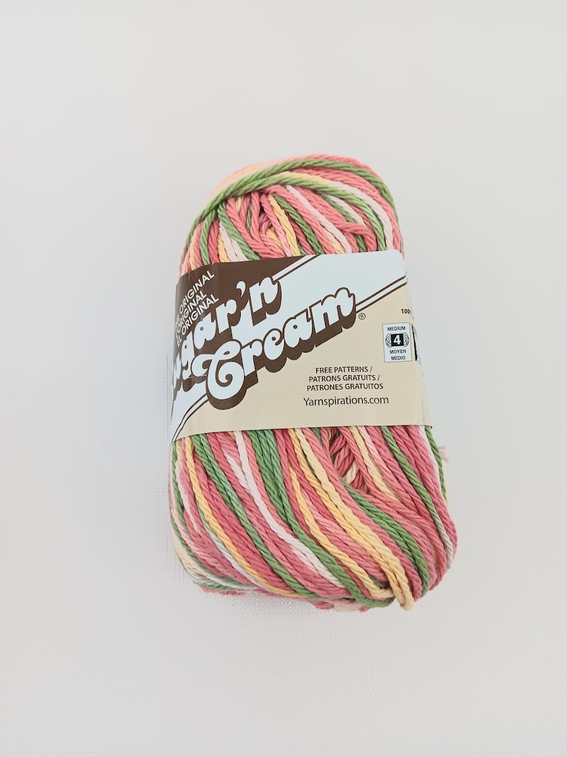 May include: A skein of Sugar'n Cream yarn featuring a variegated color palette of pink, green, yellow, and white strands. The paper label displays the brand name, a medium weight 4 symbol, and the website Yarnspirations.com for free patterns.