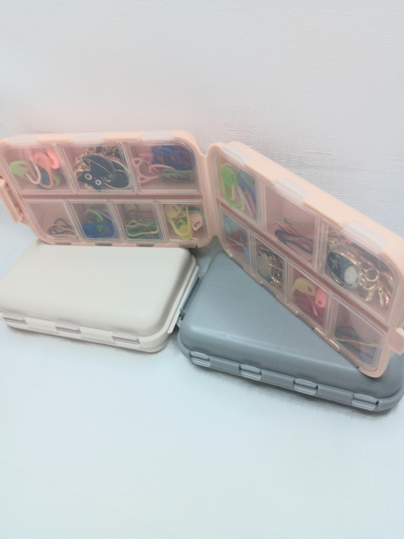 May include: Two rectangular plastic storage cases with hinged lids, one beige and one gray, each featuring multiple internal compartments filled with assorted colorful metal stitch markers, small charms, and various crafting notions.