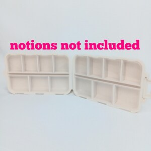 May include: Two open, off-white plastic storage containers with multiple rectangular compartments. The text "notions not included" is printed in bright pink letters above the containers. Each container features a hinged design with dividers.