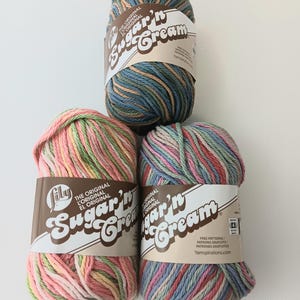 May include: Three skeins of Lily Sugar'n Cream yarn. The top skein features blue, green, and tan variegated fibers. The bottom left skein is pink, yellow, and green. The bottom right skein is a mix of pink, blue, and green. Each has a paper label.