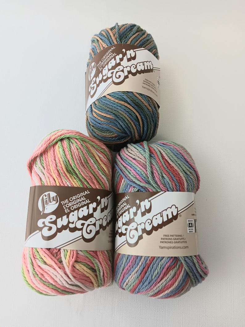 May include: Three skeins of Lily Sugar'n Cream yarn. The top skein features blue, green, and tan variegated fibers. The bottom left skein is pink, yellow, and green. The bottom right skein is a mix of pink, blue, and green. Each has a paper label.