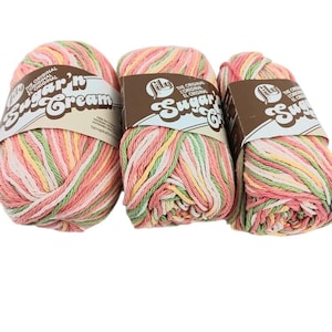 May include: Three skeins of Lily Sugar'n Cream yarn featuring a variegated color palette of soft pink, light green, and cream. Each skein is wrapped in a brown paper label with the brand name printed in white, stylized, bold lettering.