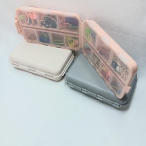 May include: Two rectangular plastic storage cases with hinged lids, one beige and one gray. Each case features multiple clear compartments filled with small colorful items, including metal charms, clips, and various small accessories.