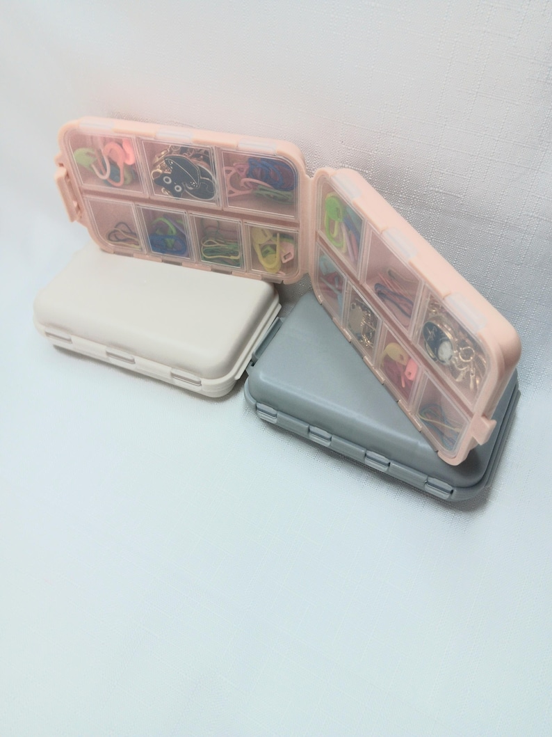 May include: Two rectangular plastic storage cases with hinged lids, one beige and one gray. Each case features multiple clear compartments filled with small colorful items, including metal charms, clips, and various small accessories.