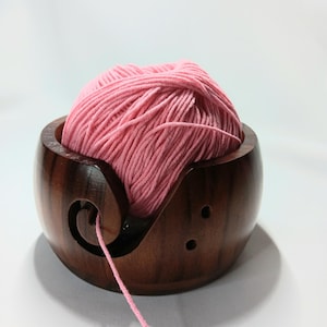 May include: A wooden yarn bowl with a pink ball of yarn inside. The yarn is threaded through the bowl's built-in yarn guide.