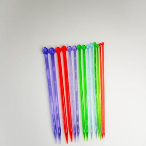 May include: A set of 11 colorful plastic crochet hooks. The hooks are arranged in a row from left to right, with the purple hooks on the left and the red hooks on the right. The hooks are all the same size and have a pointed tip.