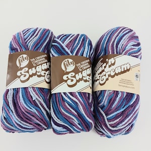 May include: Three skeins of Lily Sugar'n Cream yarn in shades of blue, purple, and white. Each skein has a brown label with the brand name and product details. The yarn is tightly wound and ready for crafting projects.