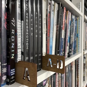 May include: A white bookshelf with a variety of DVDs and books. The DVDs are organized by letter, with a wooden divider labeled "A-D" on the top shelf. The books are mostly paperbacks and hardcovers, and they are arranged by color and size.