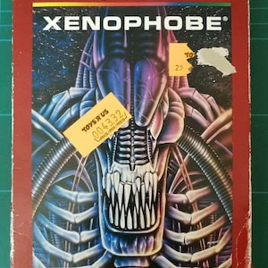 May include: A red Atari 2600 video game cartridge box featuring a black and blue alien creature on the front. The game title is "XENOPHOBE".