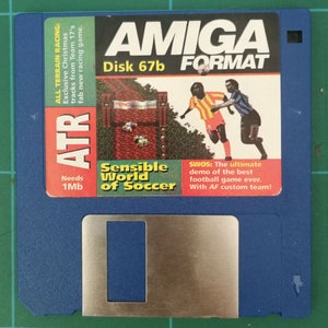 Puede incluir: Un disquete azul Amiga con una cubierta de metal plateado. La etiqueta es blanca con texto negro que dice "AMIGA FORMAT Disco 67b". La imagen muestra un juego llamado "Sensible World of Soccer" con el texto "SWOS: La demo definitiva del mejor juego de fútbol de todos los tiempos. Con el equipo personalizado AF!"