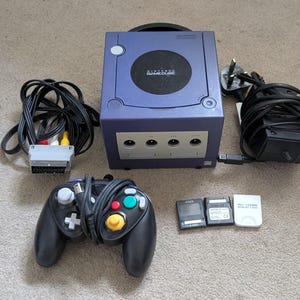 May include: A purple Nintendo GameCube console with a black controller, a power adapter, an AV cable with a SCART connector, and three memory cards. The console features a circular disc cover with the Nintendo GameCube logo printed in white text.
